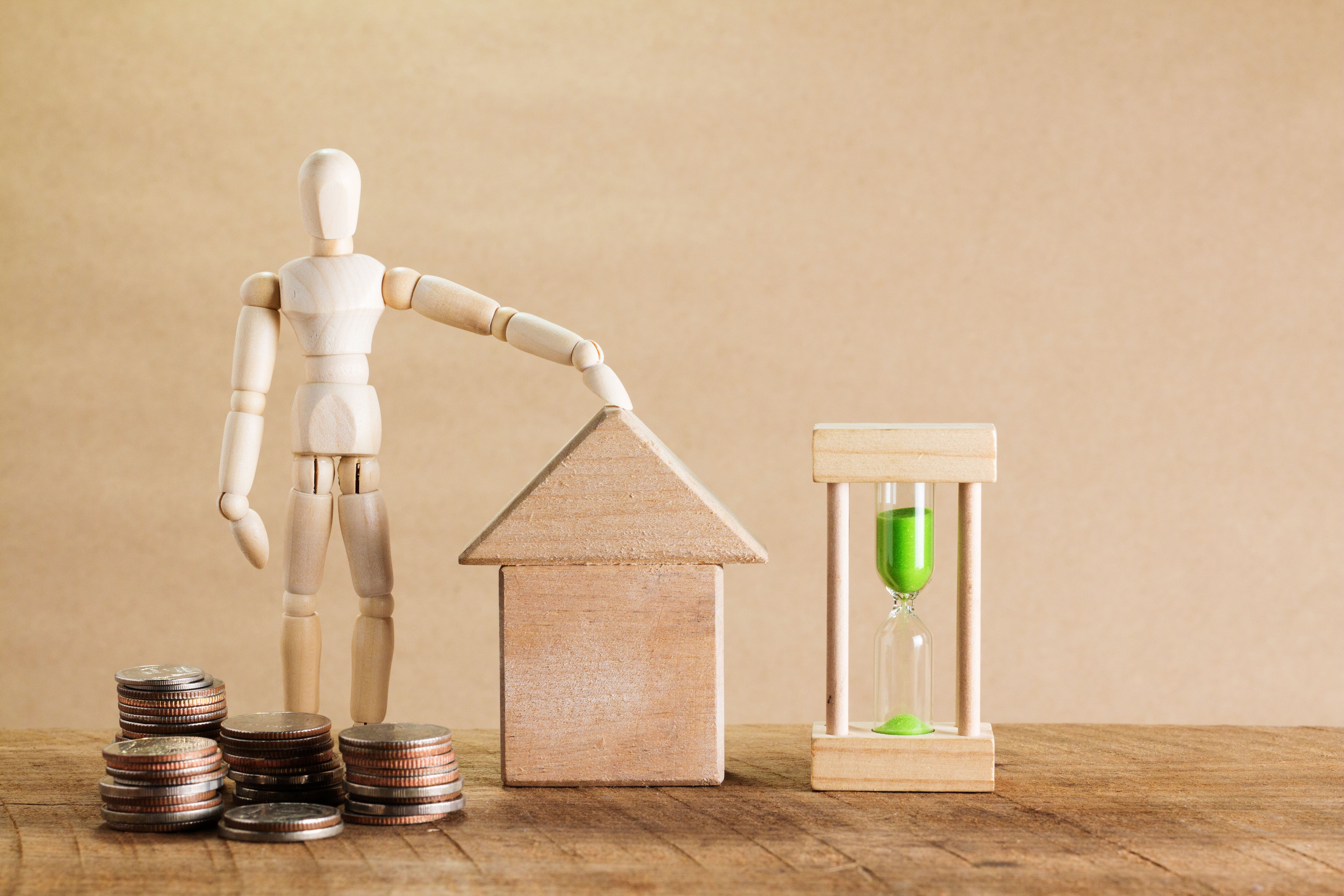 small wooden mannequin standing with a pile of coins around its feet with its left hand on a smaller house made of wooden blocks and an hourglass with green sand